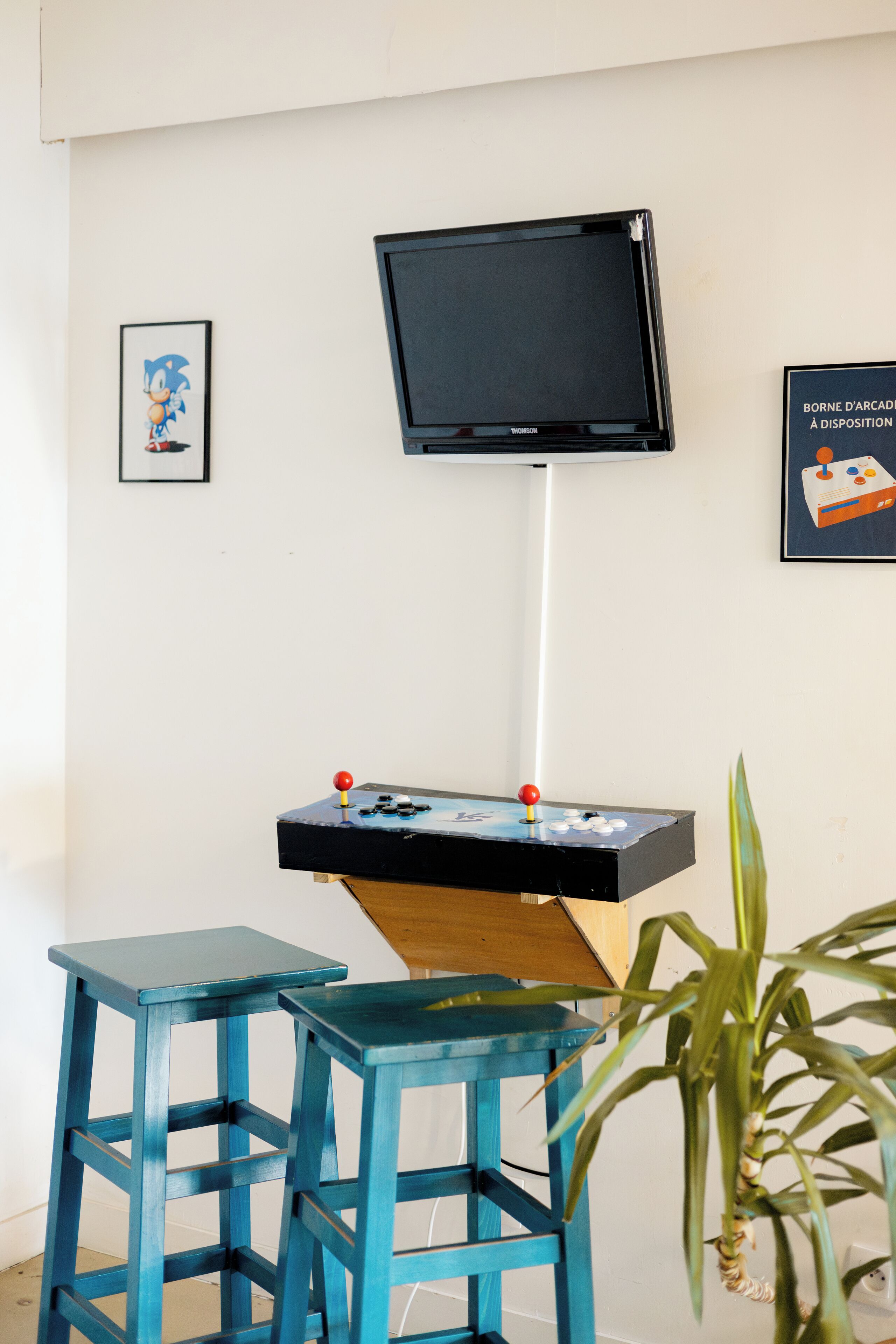 Ein Zimmer mit montiertem Fernseher, einem kleinen Tisch mit Spielautomaten-Steuerung und zwei blauen Stühlen.