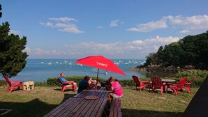 Una terraza de madera con sillas rojas y una mesa de picnic, con vistas a un mar tranquilo y barcos.