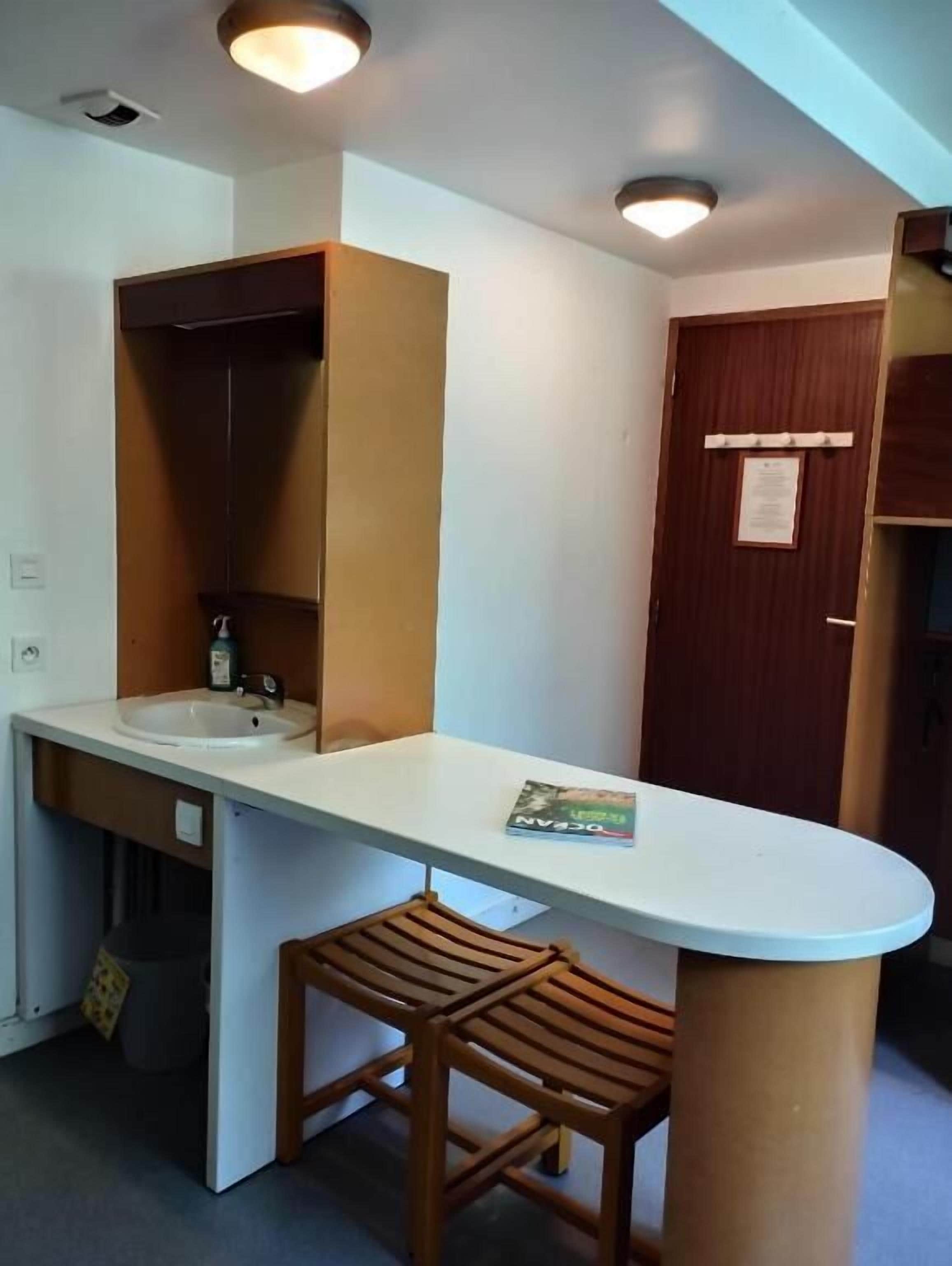 A compact kitchenette with a white countertop, a sink, and a wooden stool.