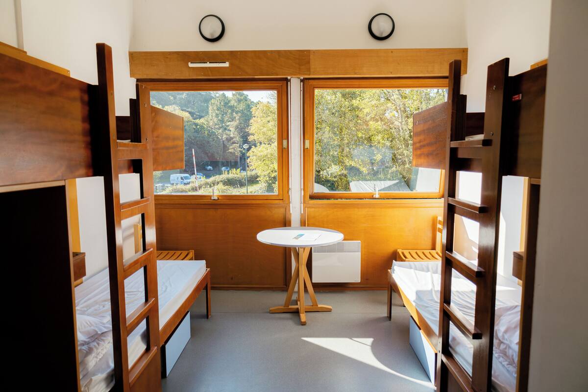 Una habitación pequeña con literas, una mesita redonda y una ventana que ofrece vistas a árboles y un edificio.