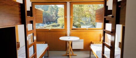 Una habitación pequeña con literas, una mesita redonda y una ventana que ofrece vistas a árboles y un edificio.
