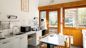 Una cocina moderna con alguien lavando platos, un comedor con mesa y sillas, y un amplio ventanal con vistas a un puente y vegetación.