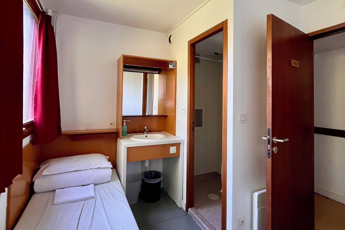Una habitación de hotel compacta con una cama individual, un baño pequeño con lavamanos y espejo, y una puerta que da a un pasillo.