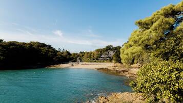 A coastal landscape with a clear blue sea, sandy beach, and a house with a sloped roof.