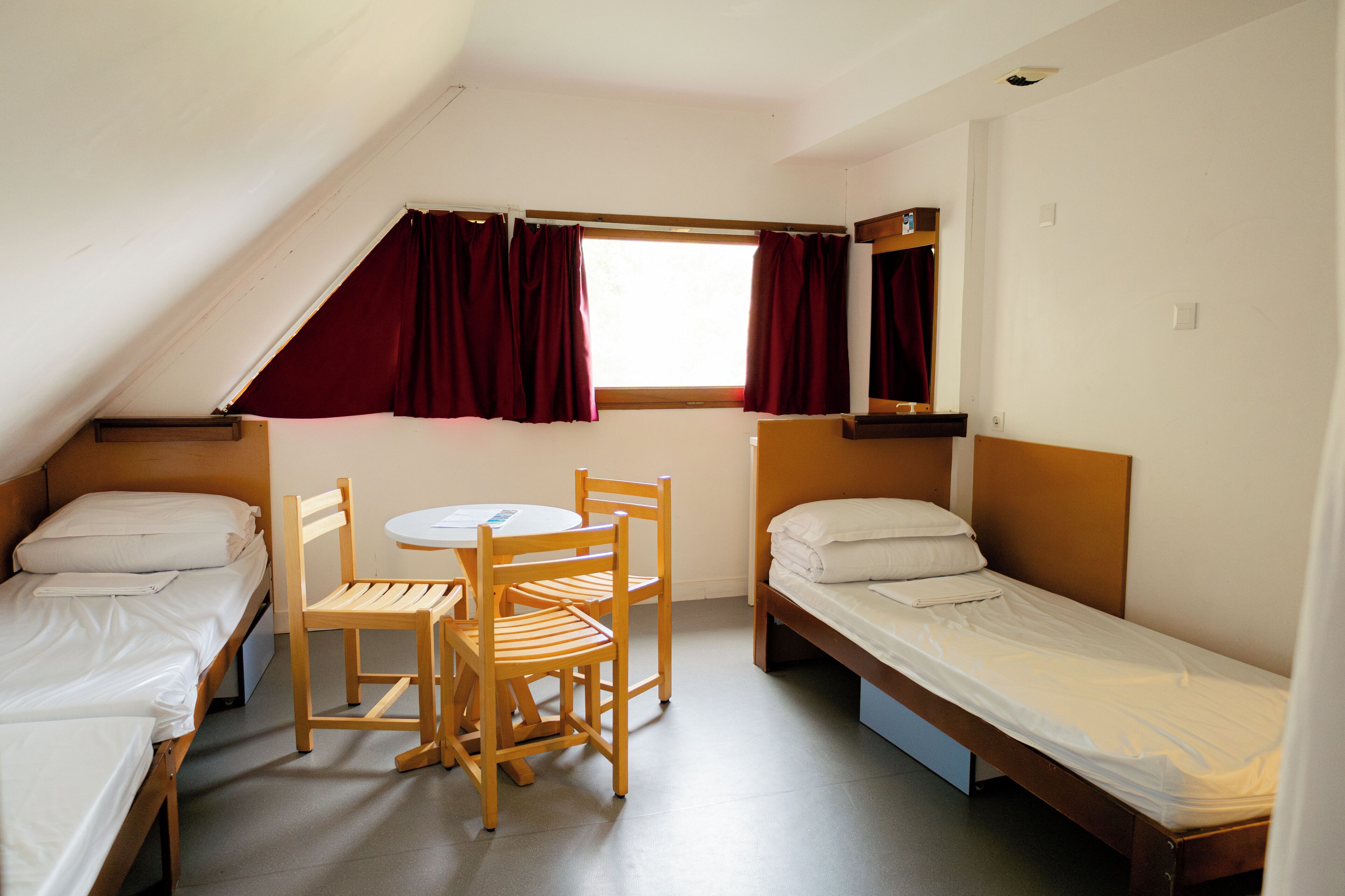 A dormitory room with two single beds, a wooden table with chairs, a window with red curtains, and a small shelf above one bed.