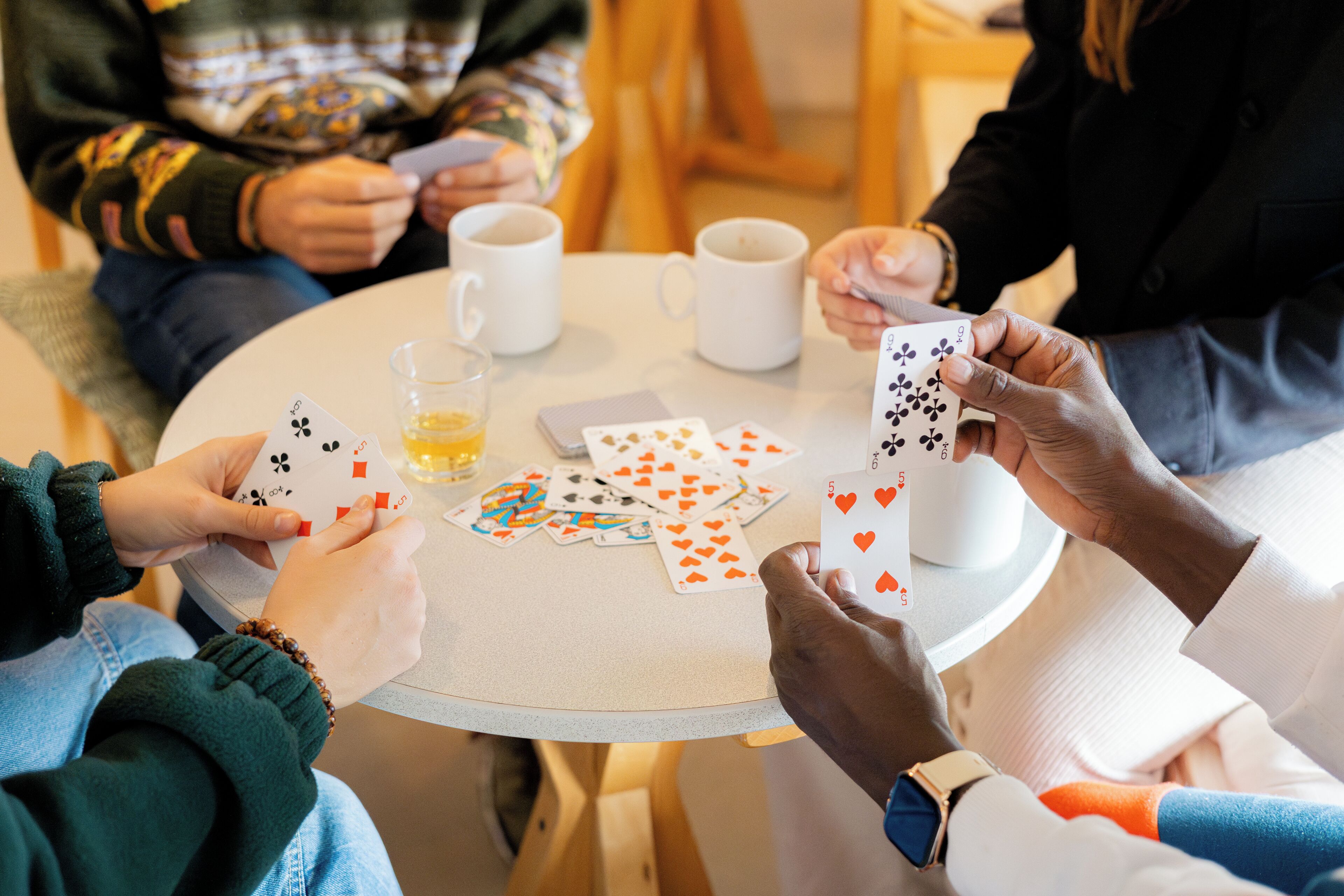 Um grupo de pessoas jogando cartas ao redor de uma mesa redonda pequena, com xícaras de café e um copo de suco de laranja.