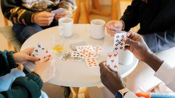 Um grupo de pessoas jogando cartas ao redor de uma mesa redonda pequena, com xícaras de café e um copo de suco de laranja.
