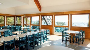 Un comedor amplio con varias mesas y sillas, grandes ventanales con vistas al mar y una estructura de techo de madera.
