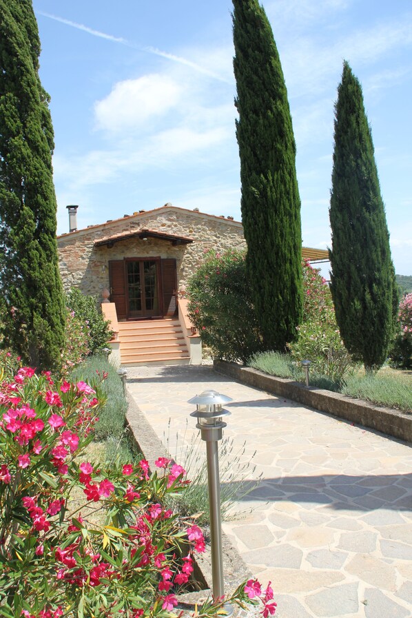 A stone building with a wooden staircase leading to a door, surrounded by tall cypress trees and pink flowers.