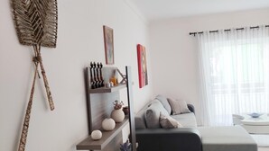 Un salon moderne avec un canapé gris, une étagère en bois ornée d’objets décoratifs, une tapisserie murale tissée et une fenêtre avec des rideaux légers.