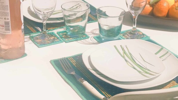 Une table est dressée pour deux personnes, avec une bouteille de vin rosé, deux verres et une assiette avec une fourchette et un couteau. On aperçoit un saladier rempli de fruits à l’arrière-plan.