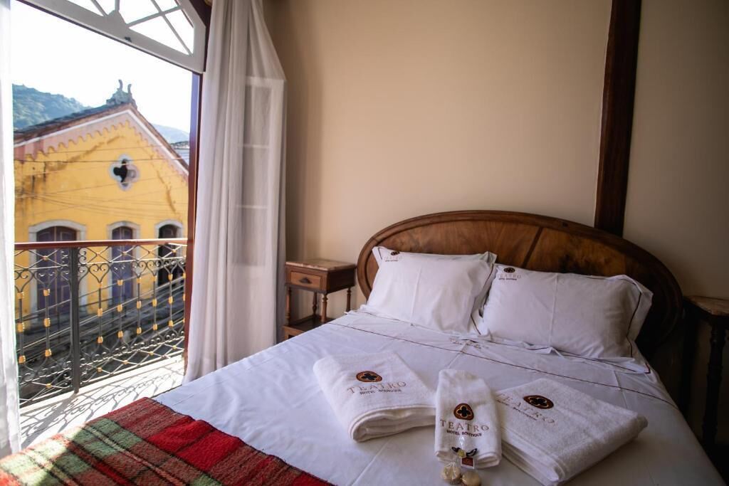 Quarto de hotel com cama, roupa de cama branca e varanda com vista para um edifício amarelo.