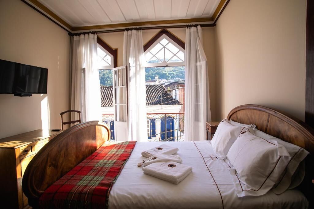 A bedroom with a bed, a red plaid blanket, white towels, and a view of a balcony with a tiled roof.