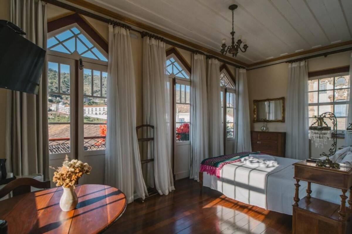 Une chambre à coucher avec un lit, un lustre, une table en bois ornée d’un vase et de grandes fenêtres avec des rideaux.