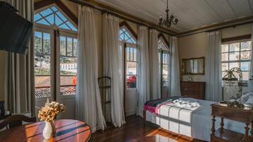 Quarto com cama, lustre, mesa de madeira com um vaso e janelas amplas com cortinas.
