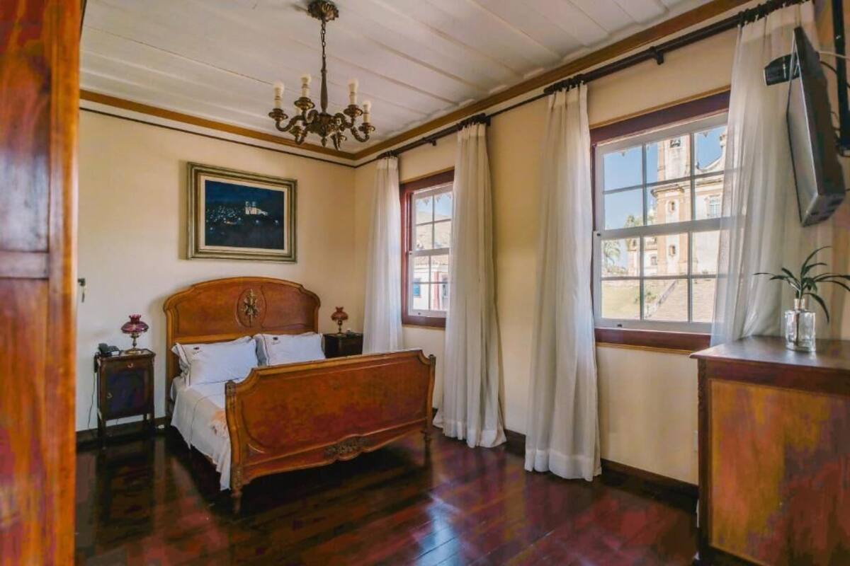 Une chambre à coucher avec un lit en bois, une table de chevet, un lustre, une fenêtre avec des rideaux et un tableau encadré au mur.