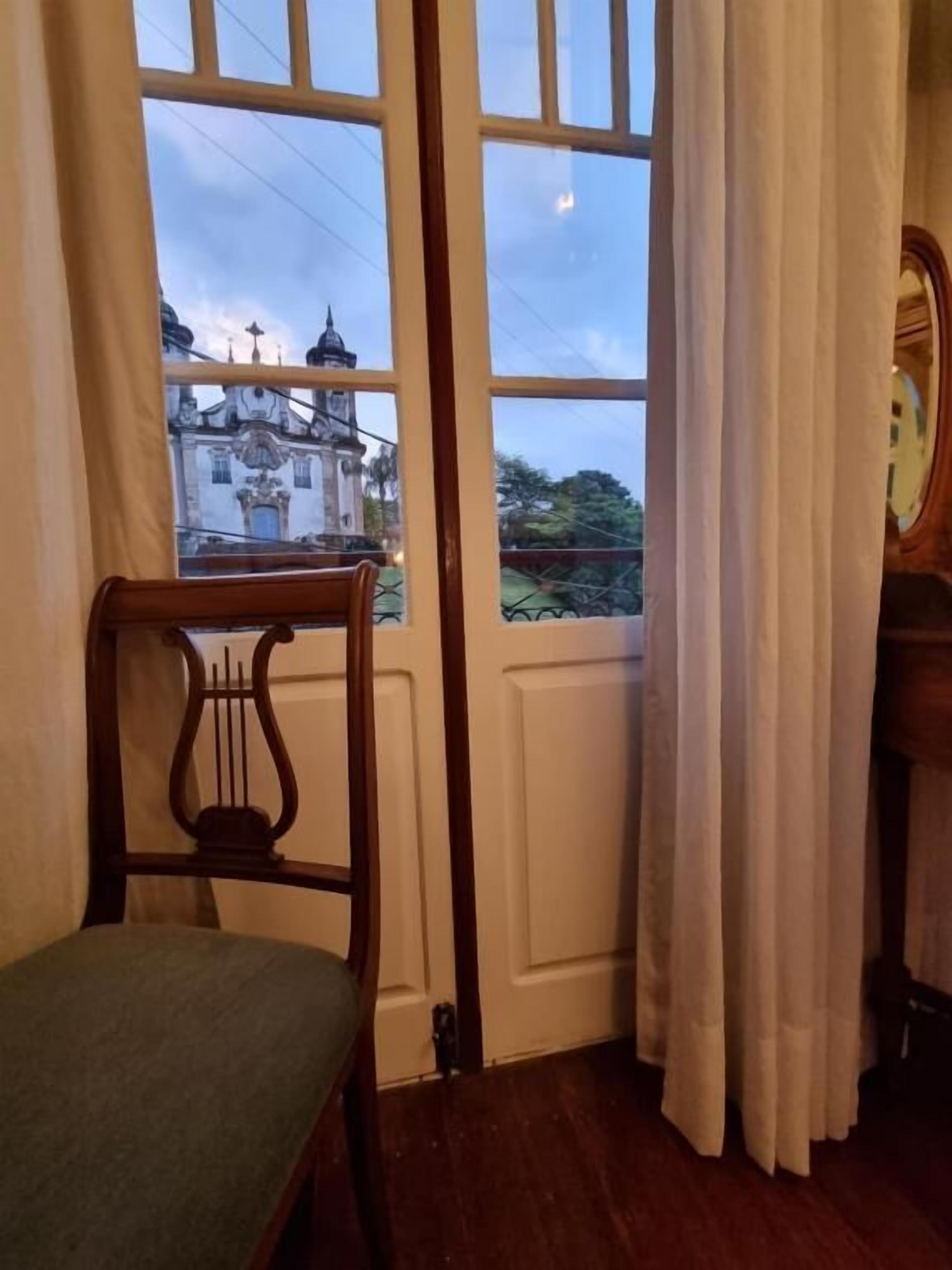 A room with a wooden chair, a piano, and a view of a building through a window.
