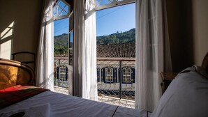 Quarto com cama, janela com cortinas e vista para um edifício com telhado ladrilhado.
