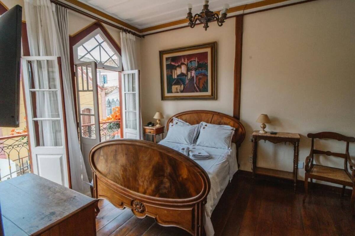 Une chambre à coucher avec un lit en bois, un tableau au mur, une fenêtre avec des rideaux et une vue sur les bâtiments à l’extérieur.