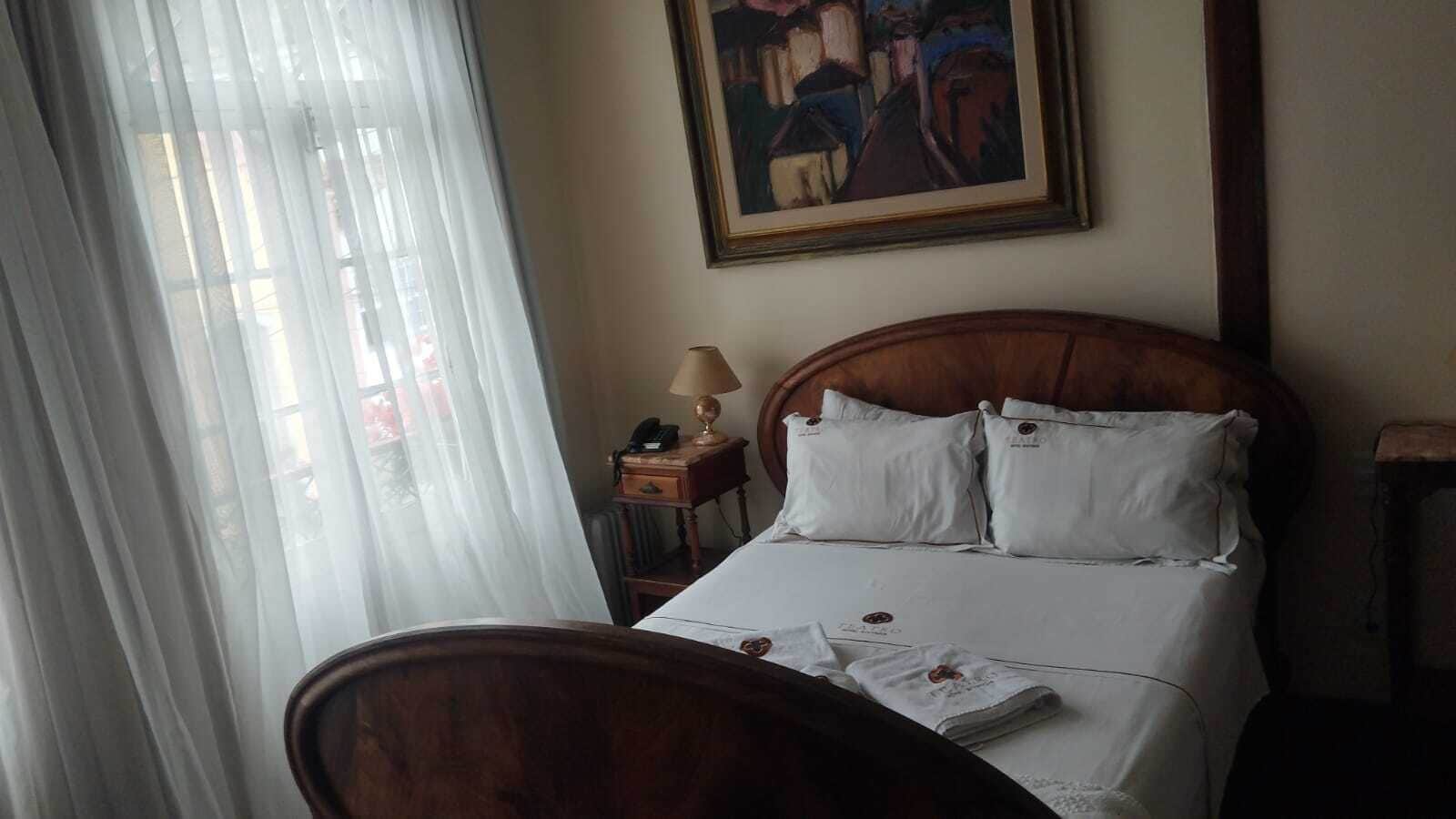 A hotel room with a bed, a desk with a lamp, a chair, a painting on the wall, and sheer curtains.