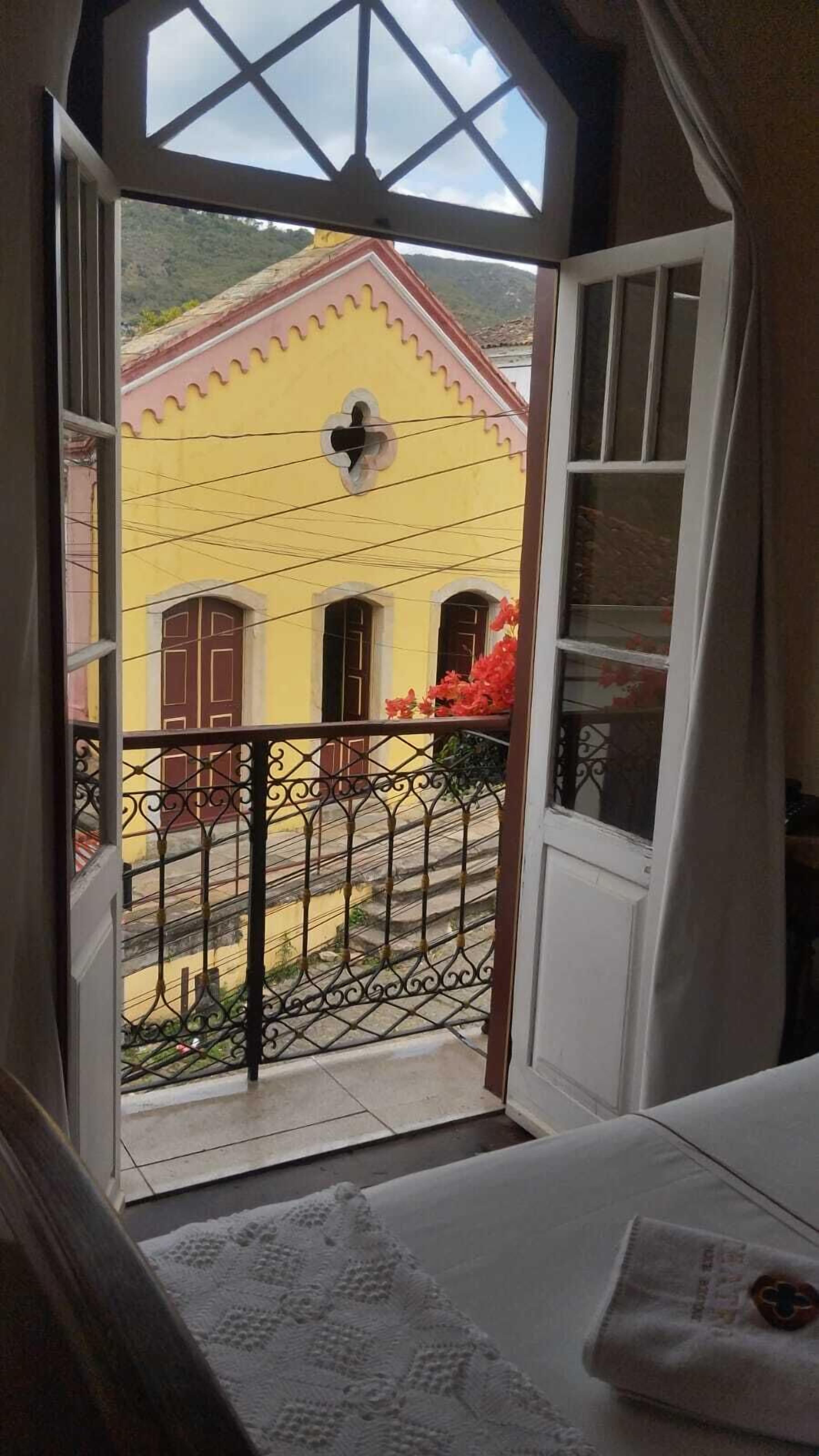 Quarto com varanda, cama e janela com vista para um edifício amarelo.