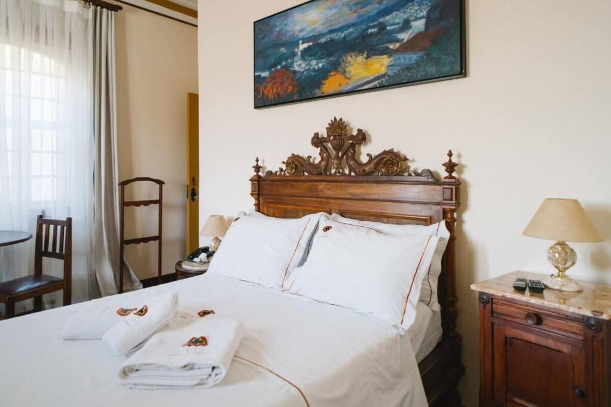 Une chambre d’hôtel avec un lit en bois, du linge de lit blanc, un tableau au mur, un bureau avec une lampe et une chaise.