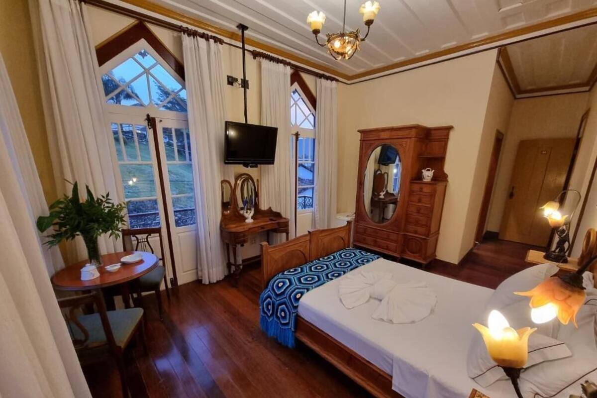 Une chambre à coucher avec un grand lit, une télévision, une commode en bois, un bureau avec une chaise et une fenêtre avec des rideaux.