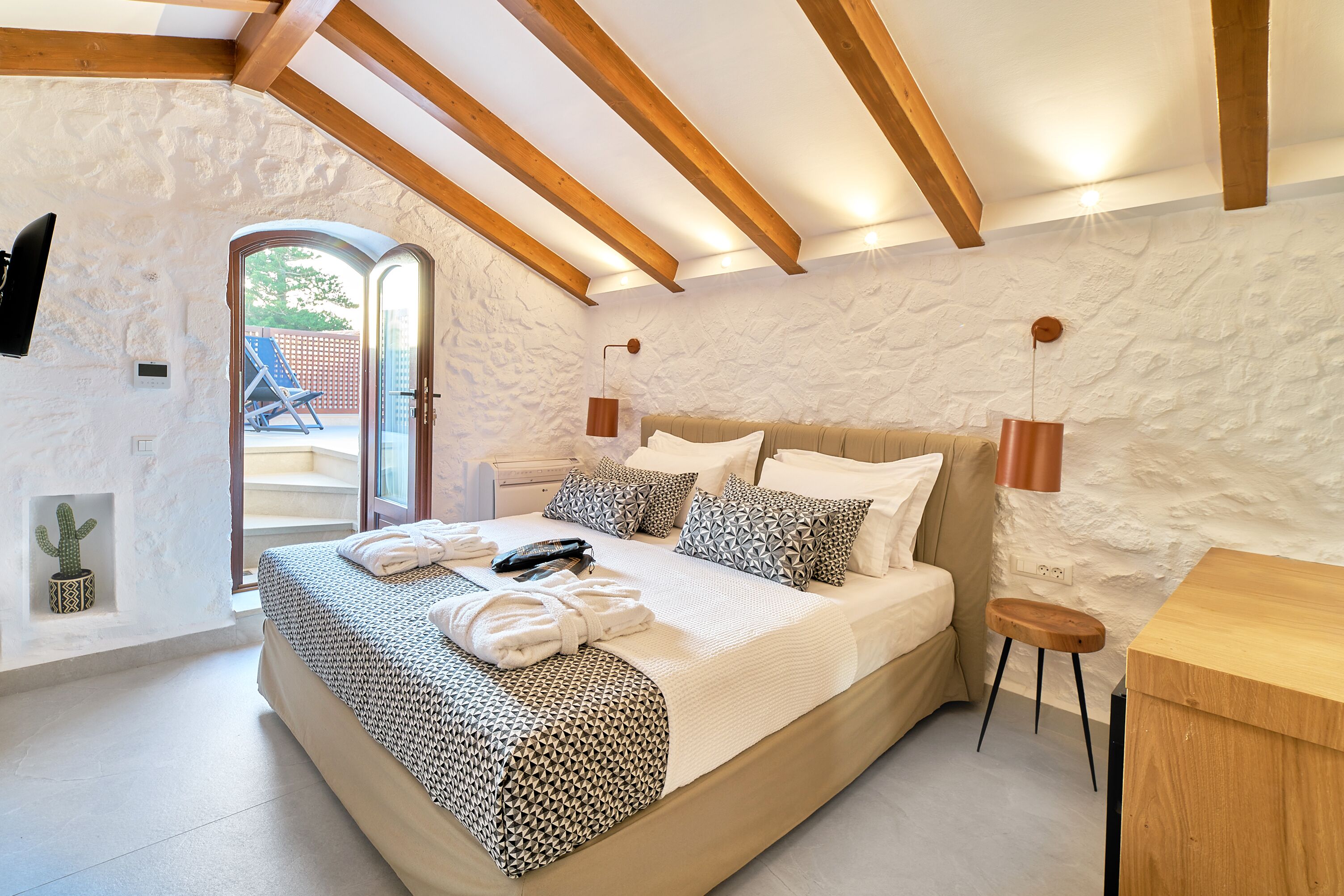 A bedroom with a large bed, wooden beams, a stone wall, a small table, and a cactus plant.