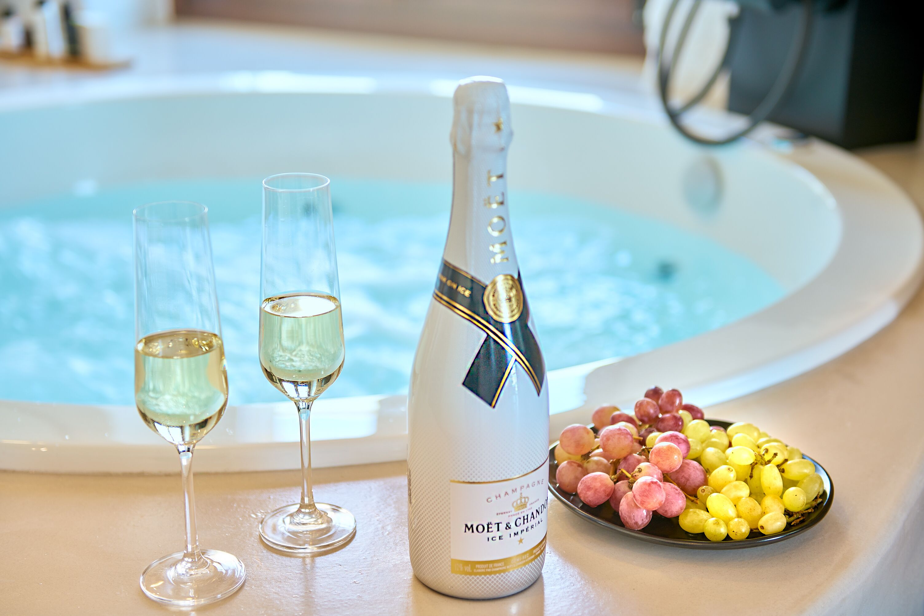 Una botella de Moet & Chandon, dos copas de champán y un bol de uvas sobre una mesa cerca de una bañera.