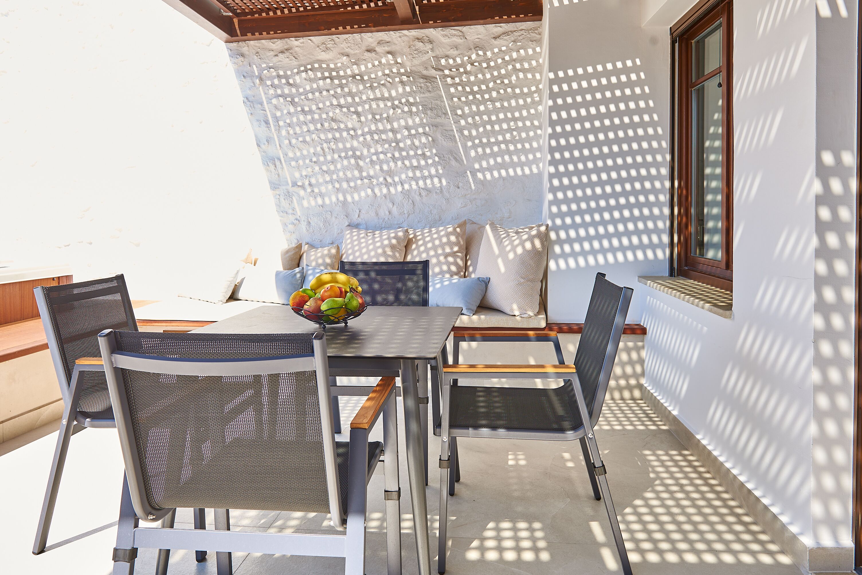 An outdoor patio with a table and chairs, a bowl of fruit, and a white wall with a lattice design.