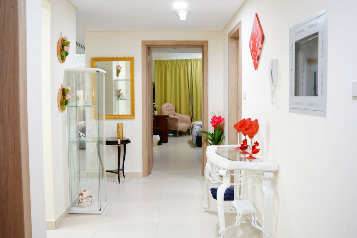 Un couloir avec une vitrine, une table blanche ornée d’un vase de fleurs rouges et un miroir accroché au mur.