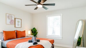 Une chambre à coucher comprenant un lit, un ventilateur de plafond, une fenêtre avec des stores, une table de chevet avec une lampe et un miroir.