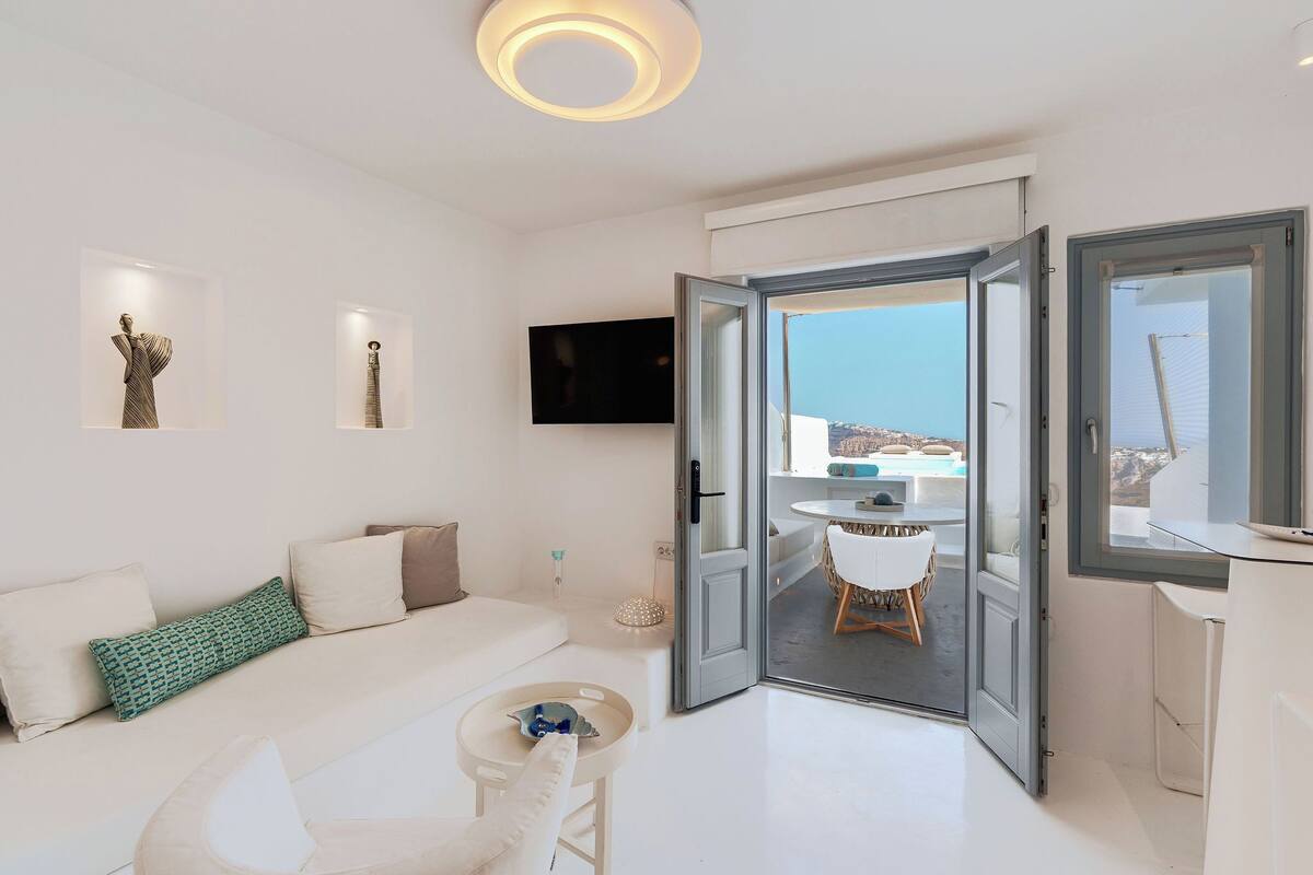 A modern, minimalist living room with a sofa, a coffee table, and a dining area with a table and chairs. A TV is mounted on the wall, and there is a balcony with a view of the sea.