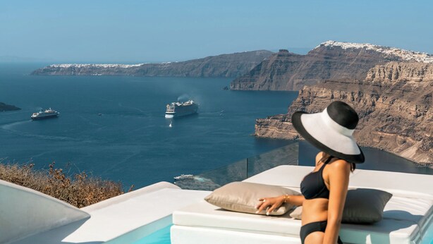 A woman in a black bikini and wide-brimmed hat sitting on a white lounge chair by a pool with clear blue water, overlooking a rocky coastline with boats in the distance. - Nelya Suites (Santorini)