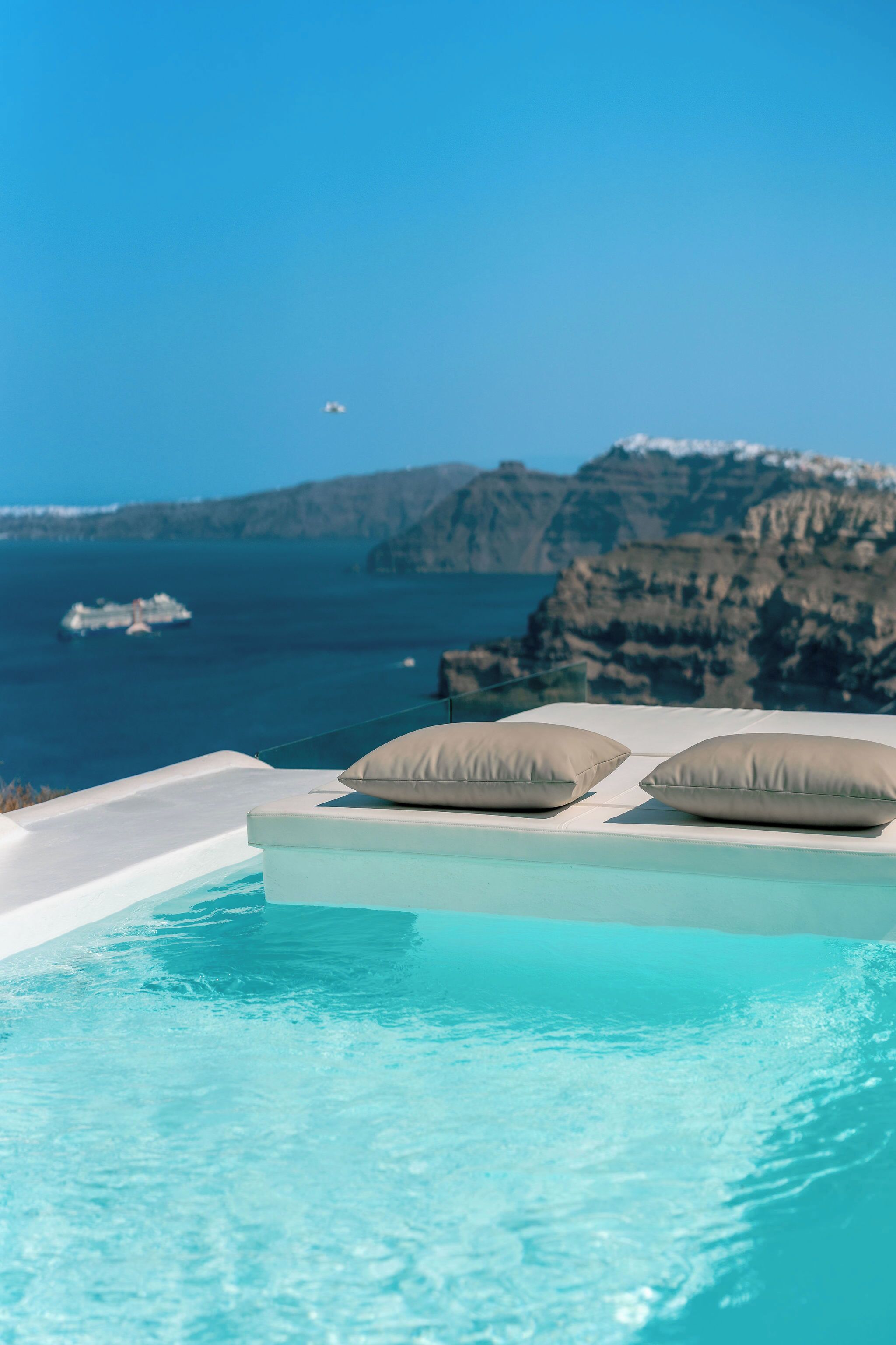 An infinity pool with two beige pillows, a clear blue sky, and a view of the sea and cliffs.