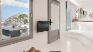 A modern hotel room with a large window offering a view of a rocky landscape, a flat-screen TV mounted on the wall, and a wooden coffee table with a decorative item on it. - Nelya Suites (Santorini)