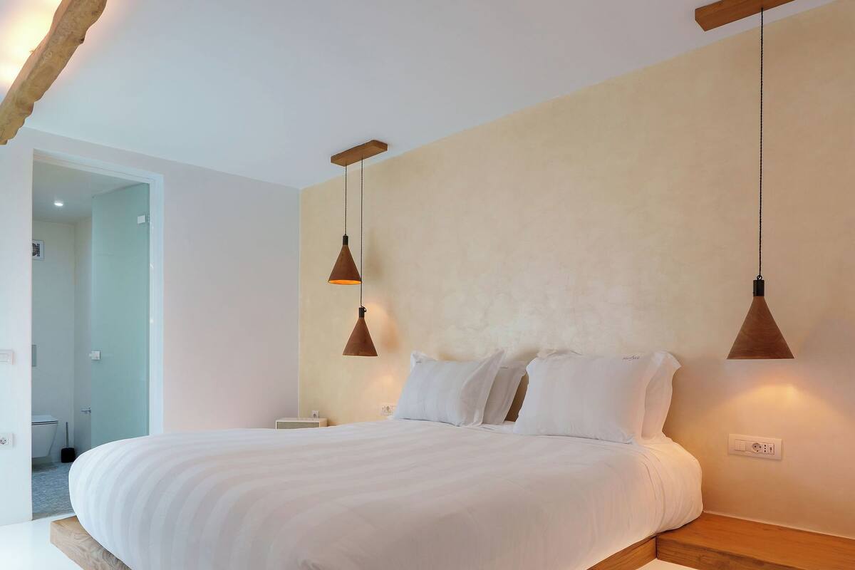 A modern bedroom with a large bed, wooden headboard, and pendant lighting.