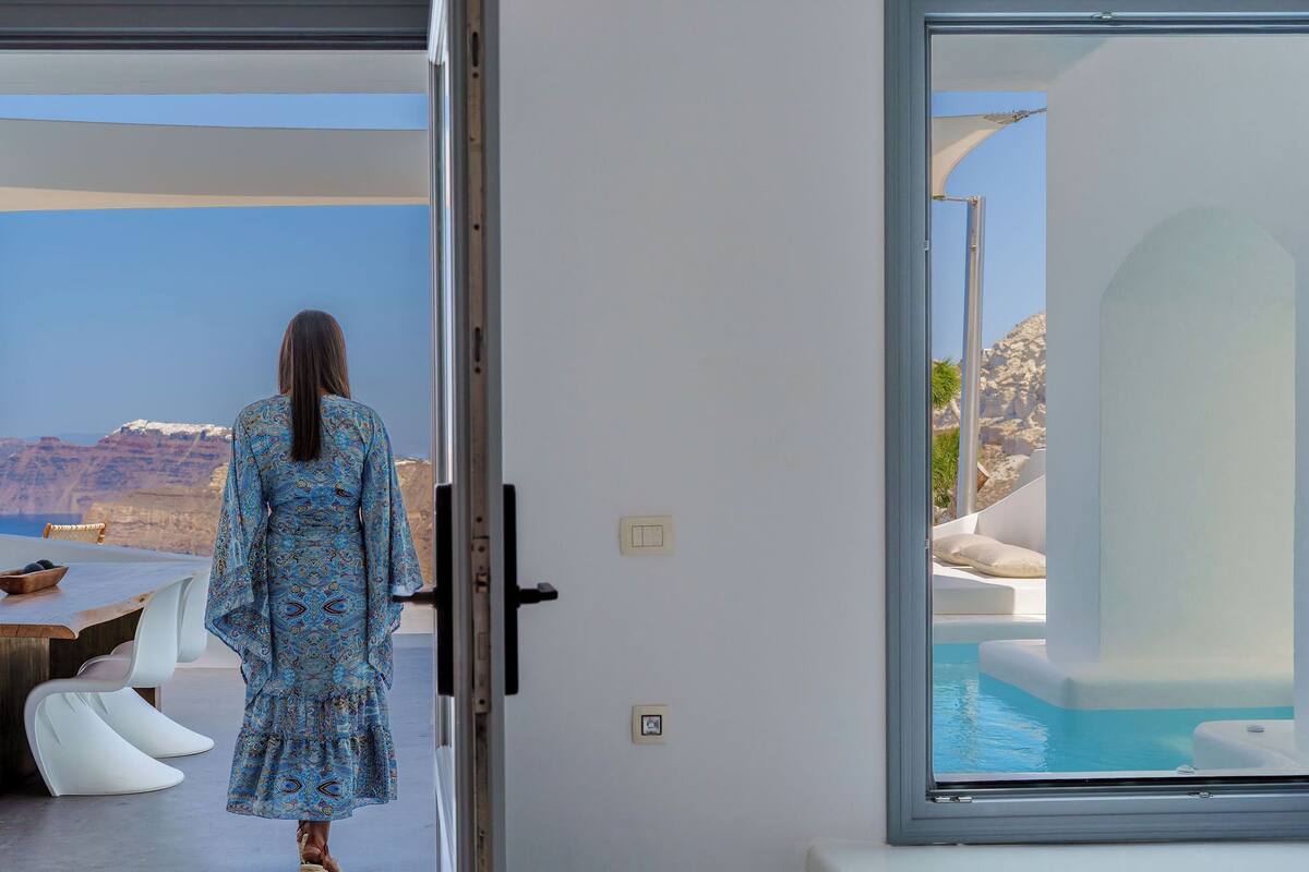 A woman in a patterned dress standing by a sliding glass door, overlooking a pool and a mountainous landscape.