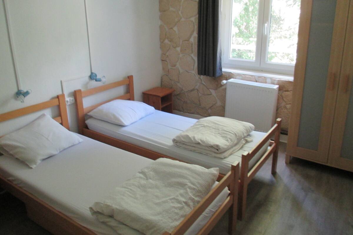 Zwei Einzelbetten mit Holzrahmen und weißen Bettwäsche, eine Steinwand, ein Heizkörper und ein Fenster mit Vorhängen.