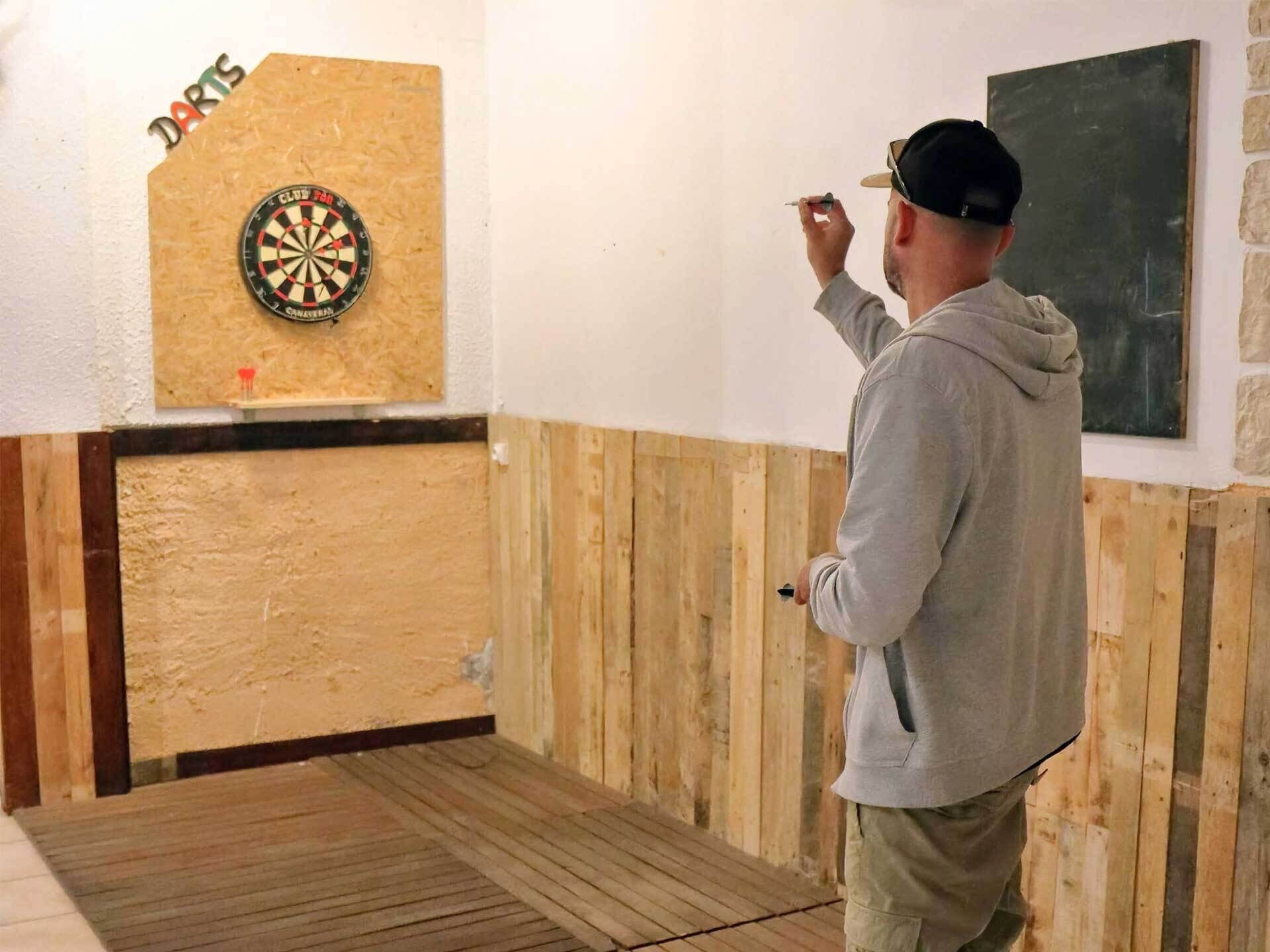 Eine Person spielt Darts in einem Raum mit Holzvertäfelung und einem Dartsbrett an der Wand.