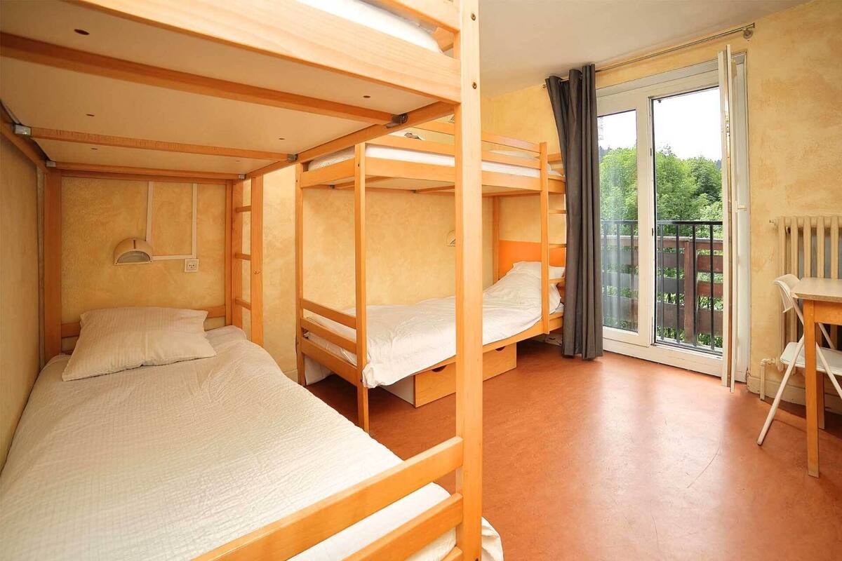 Ein Schlafsaalzimmer mit drei Etagenbetten, einem kleinen Tisch und einem Fenster mit Blick auf Grünfläche.
