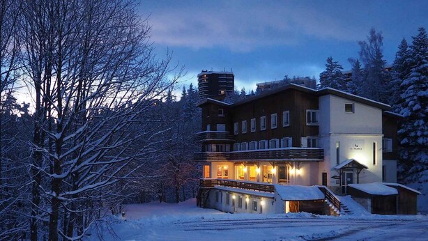 Un bâtiment recouvert de neige, avec un escalier menant à l’entrée, entouré d’arbres et sous un ciel dégagé.
