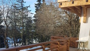 Une personne est assise à une table, un repas devant elle, sur une terrasse en bois, avec vue sur des arbres et un bâtiment en arrière-plan.