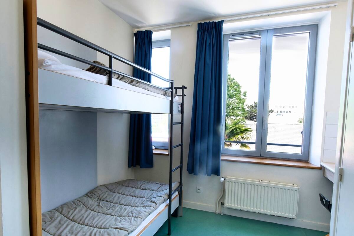 A dormitory room with bunk beds, a window with curtains, and a radiator.