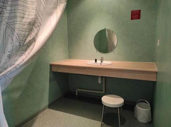 A small bathroom with a single sink, a mirror, a stool, and a caution sign on the wall. - Auberge de Jeunesse HI Cherbourg (Cherbourg-en-Cotentin)