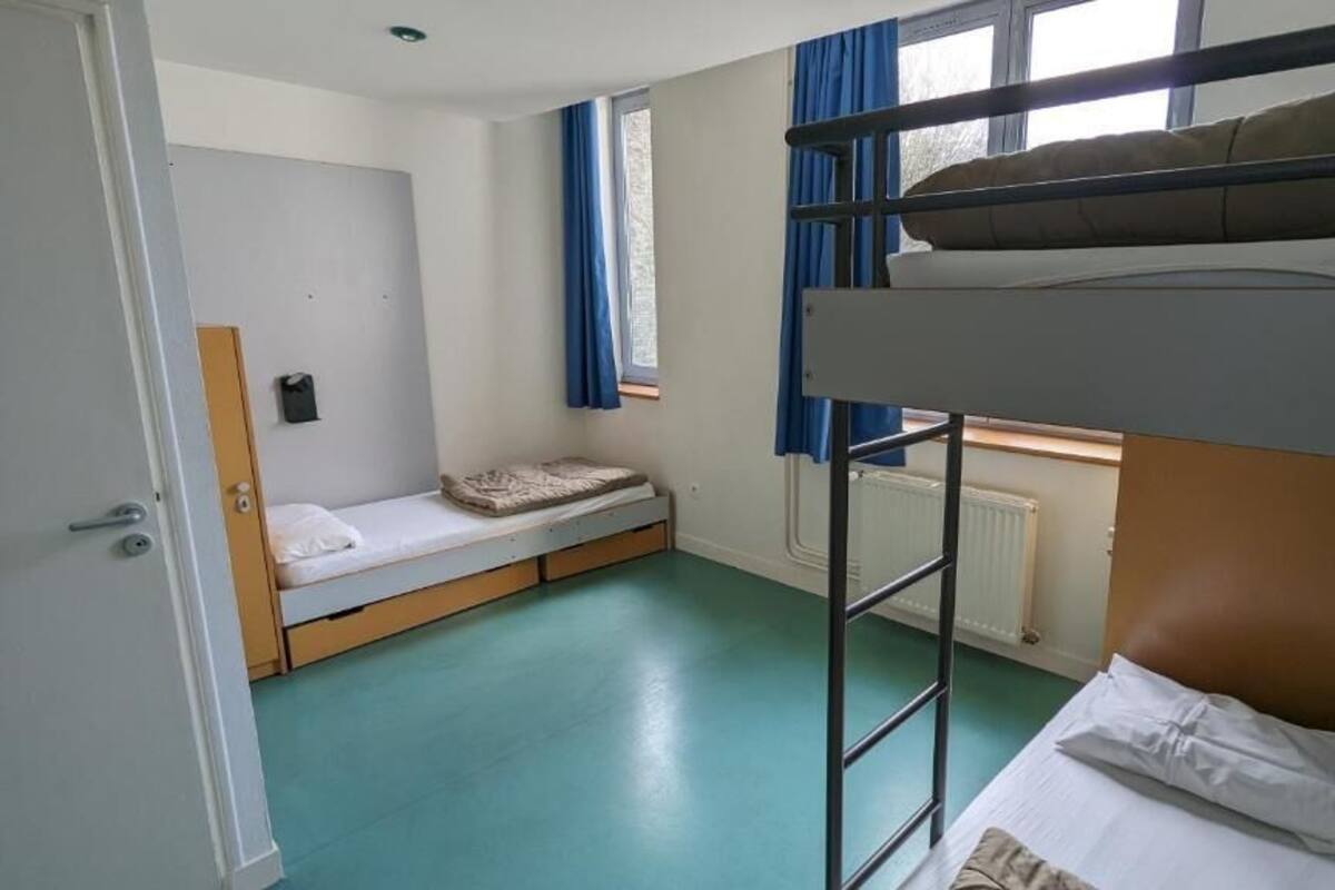 A dormitory room with bunk beds, a small window with curtains, and a radiator.
