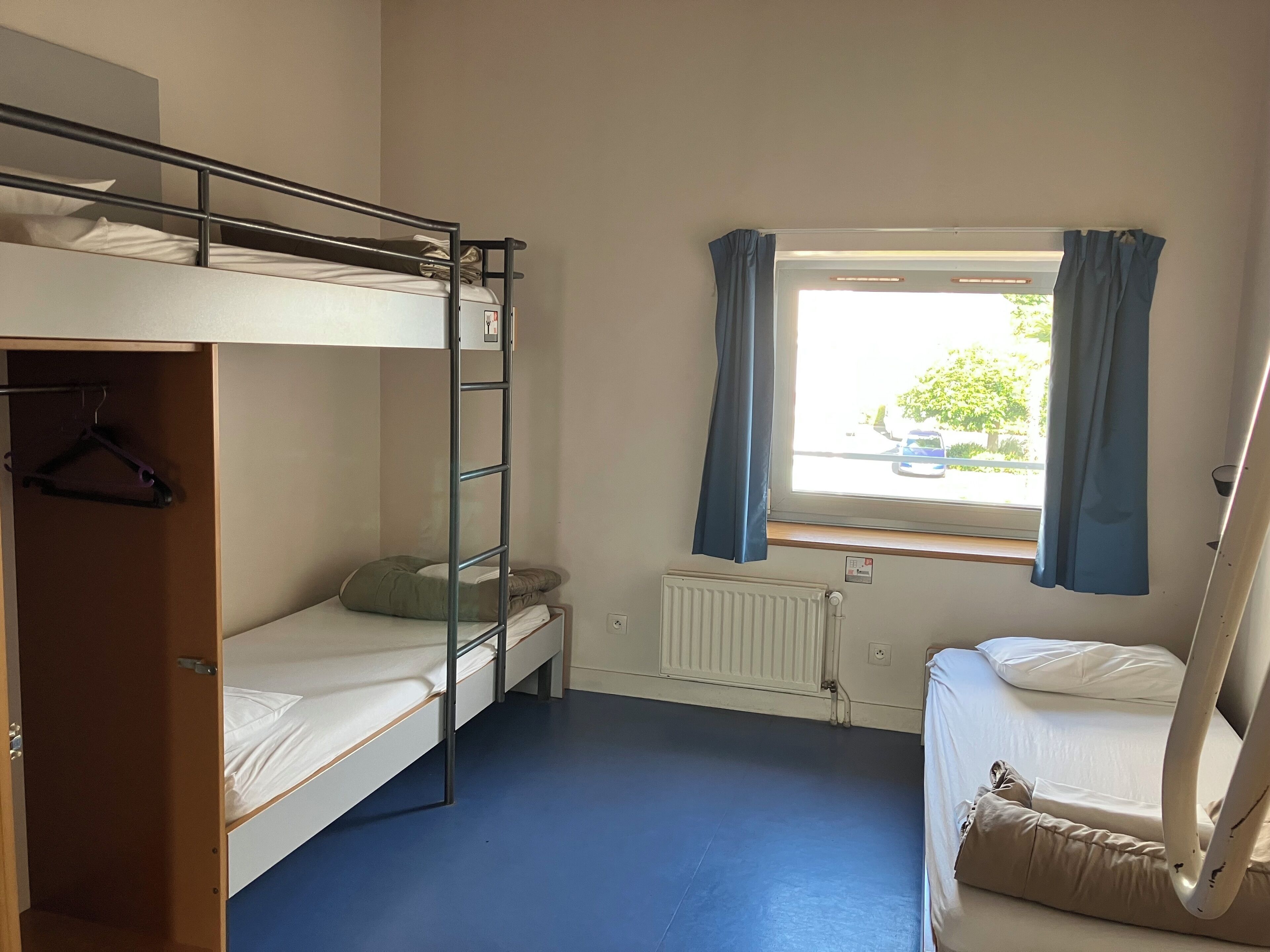 Un dormitorio con literas, una ventana con cortinas, un radiador y una puerta con perchero.