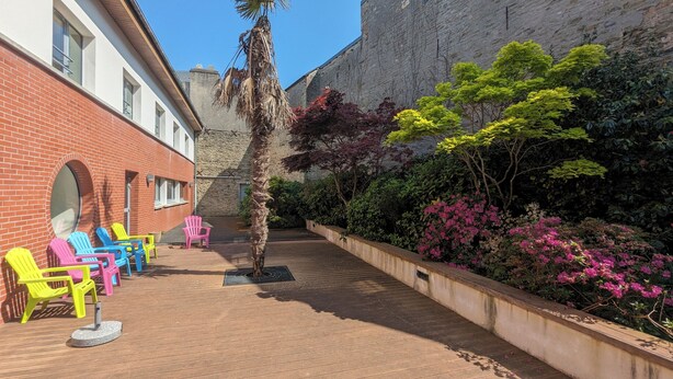 A courtyard with colorful chairs, a palm tree, and a variety of flowering plants. - Auberge de Jeunesse HI Cherbourg (Cherbourg-en-Cotentin)