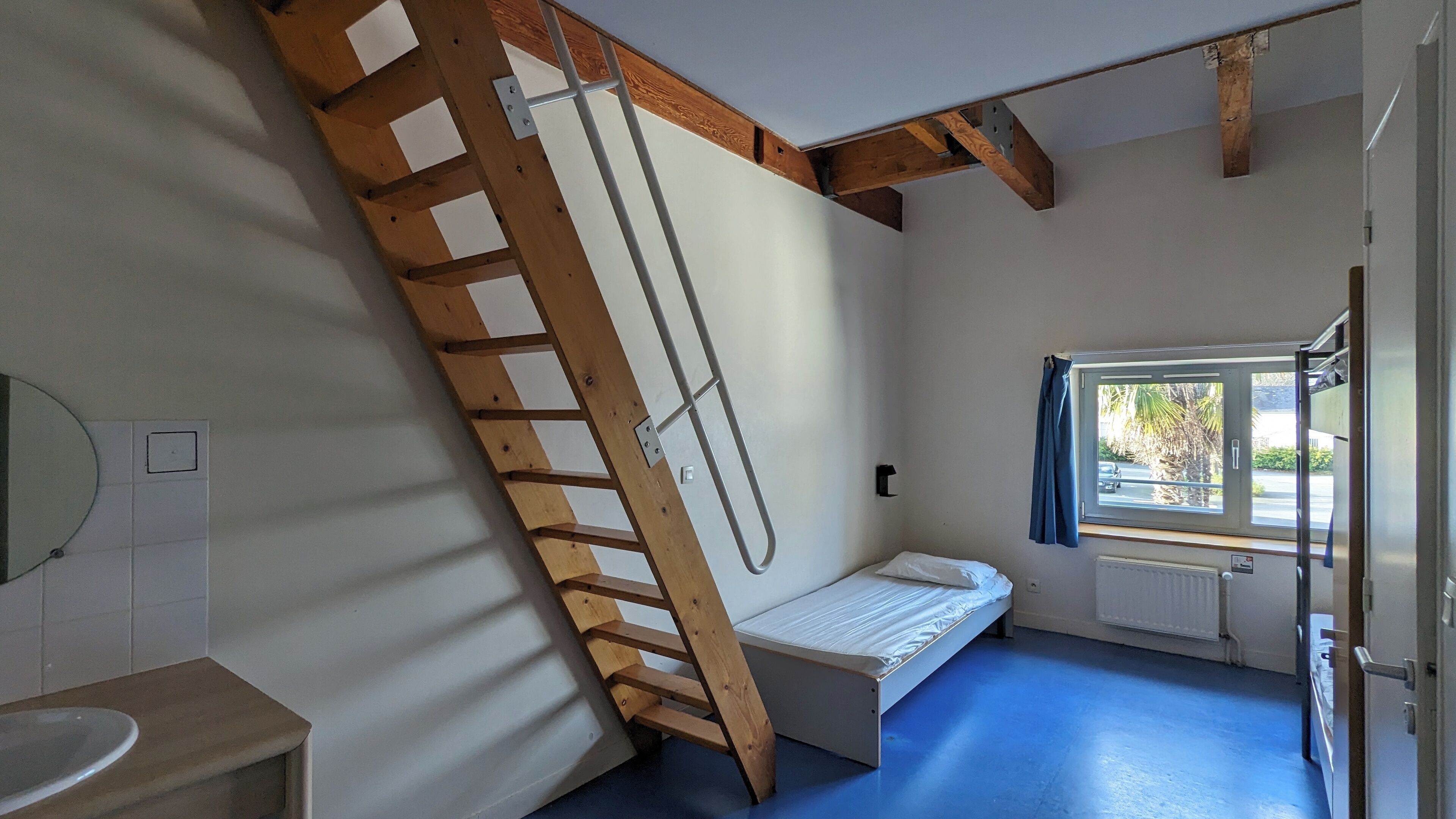 A room with a wooden ladder, a bed, a window with curtains, a radiator, and a sink.