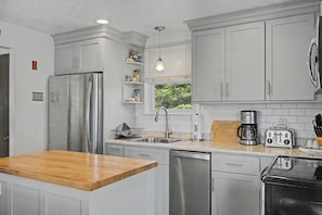 Uma cozinha com armários brancos, eletrodomésticos de aço inoxidável, bancada de madeira e uma janela com persianas.